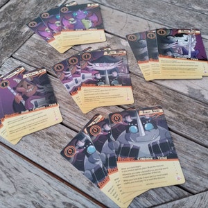 Pode incluir: Um conjunto de cartas de jogo coloridas com personagens de desenhos animados com diferentes habilidades. As cartas estão dispostas sobre uma mesa de madeira. As cartas têm um esquema de cores roxo, amarelo e laranja. As cartas têm texto que diz "Resource: Companion - Virtual" e "Resource: Synth - Common - Virtual".