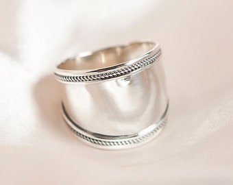 Könnte beinhalten: Ein silberner Ring mit einem breiten Band und einem strukturierten Rand. Der Ring liegt auf einem weißen Hintergrund.