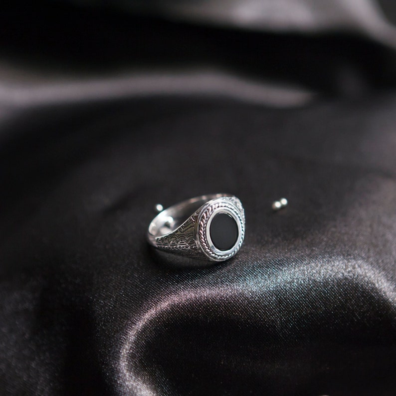 May include: A silver signet ring with a black onyx stone set in a circular bezel. The ring has an intricate engraved design around the stone.