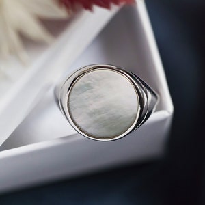 May include: A silver ring with a white oval-shaped gemstone. The ring is sitting in a white box.