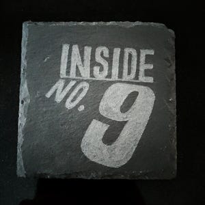 Puede incluir: Posavasos cuadrado de pizarra gris oscuro con las palabras "INSIDE NO. 9" en letras blancas. El número 9 es grande y estilizado. El posavasos tiene un borde rugoso y natural y probablemente se usa para bebidas.