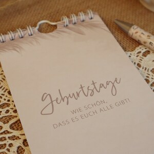 Könnte beinhalten: Ein beigefarbener Spiralblock mit dem Text "Geburtstage" in brauner Kursivschrift. Der Text darunter lautet "Wie schön, dass es euch alle gibt!"