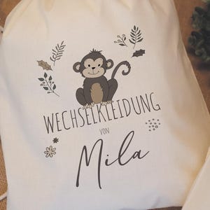 Könnte beinhalten: Ein cremefarbener Kordelzugbeutel mit einer Cartoon-Affen-Illustration, umgeben von botanischen Elementen. Der Beutel trägt den Text "WECHSELKLEIDUNG VON Mila". Der Affe ist braun und grau.