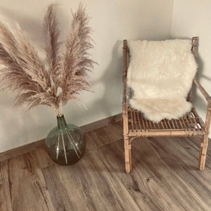 Puede incluir: Una silla de mimbre marr&oacute;n claro con un coj&iacute;n de piel de oveja blanca en el asiento. Un jarr&oacute;n con hierba de la pampa seca est&aacute; en el suelo de madera.