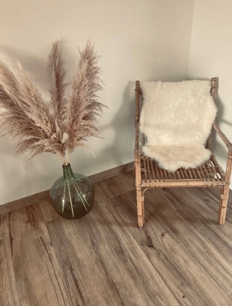 Puede incluir: Una silla de mimbre marr&oacute;n claro con un coj&iacute;n de piel de oveja blanca en el asiento. Un jarr&oacute;n con hierba de la pampa seca est&aacute; en el suelo de madera.