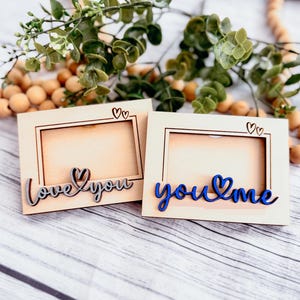May include: Two small wooden picture frames with a cutout heart design. The frame on the left says "love you" in cursive with a heart in the middle. The frame on the right says "you & me" in cursive with a heart in the middle.