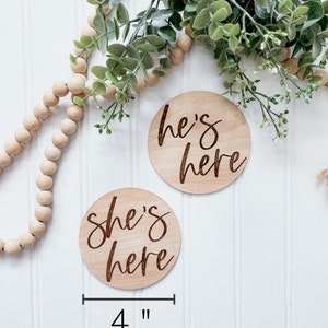 Puede incluir: Dos letreros redondos de madera de 10 cm de di&aacute;metro con el texto "She's here" y "He's here" en escritura cursiva. Los letreros est&aacute;n sobre un fondo de madera blanca.