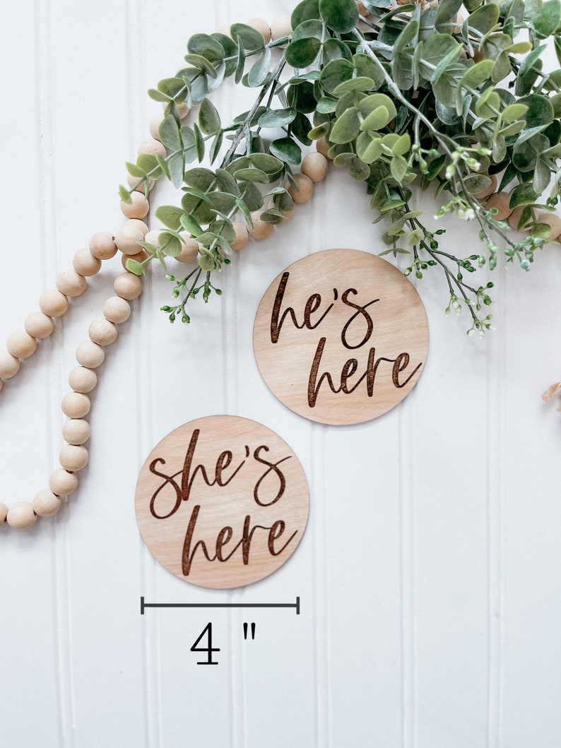 Puede incluir: Dos letreros redondos de madera de 10 cm de di&aacute;metro con el texto "She's here" y "He's here" en escritura cursiva. Los letreros est&aacute;n sobre un fondo de madera blanca.