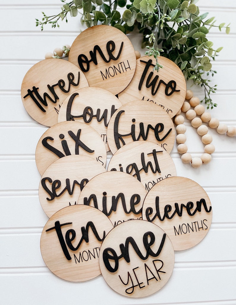 May include: A set of twelve wooden baby milestone discs with black script. The discs feature the numbers one through twelve, with the number twelve reading "one year".