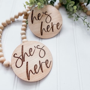 Puede incluir: Dos letreros redondos de madera con el texto "She's here" y "He's here" en una fuente cursiva. Los letreros est&aacute;n sobre un fondo de madera blanca con una cuerda de cuentas de madera.