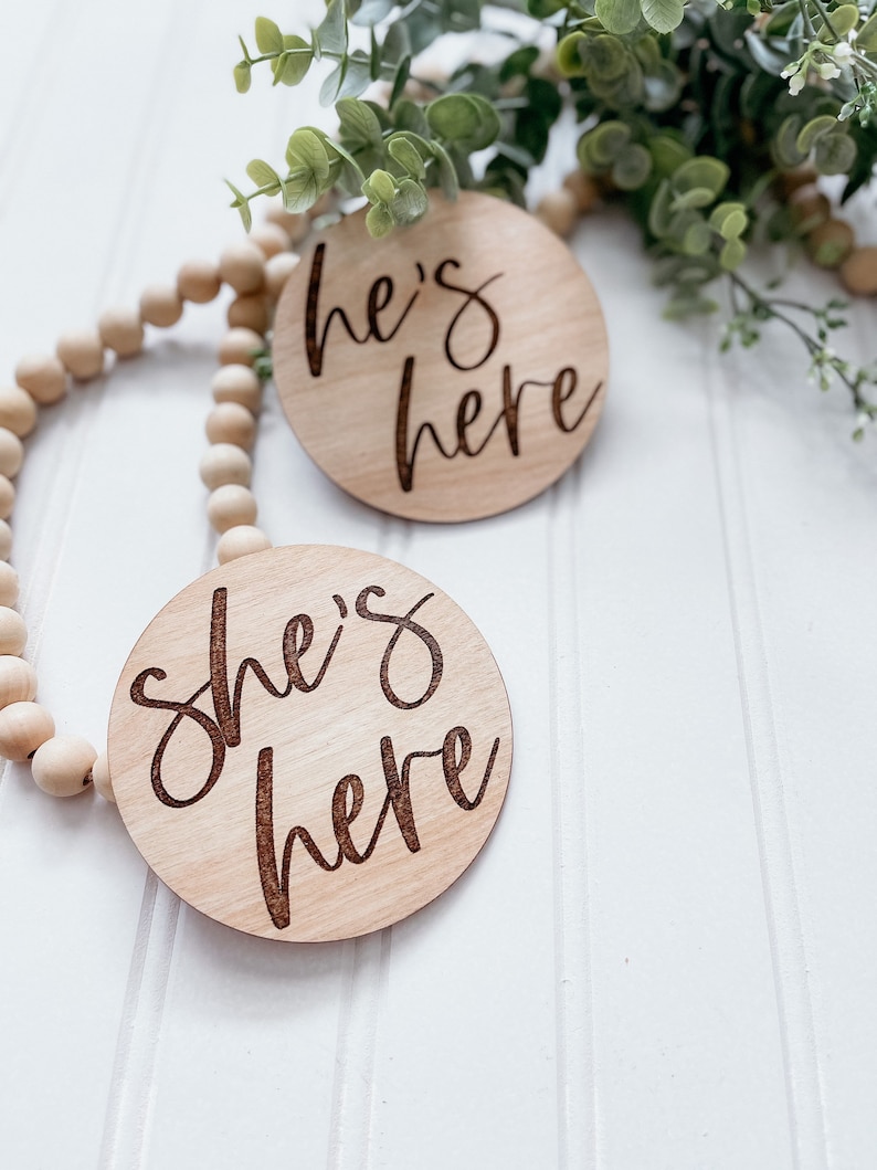 Puede incluir: Dos letreros redondos de madera con el texto "She's here" y "He's here" en una fuente cursiva. Los letreros est&aacute;n sobre un fondo de madera blanca con una cuerda de cuentas de madera.