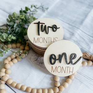 May include: Two wooden baby milestone discs with black lettering. The top disc says "two months" and the bottom disc says "one month".