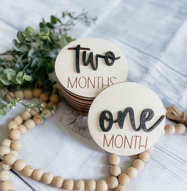 May include: Two wooden baby milestone discs with black lettering. The top disc says "two months" and the bottom disc says "one month".