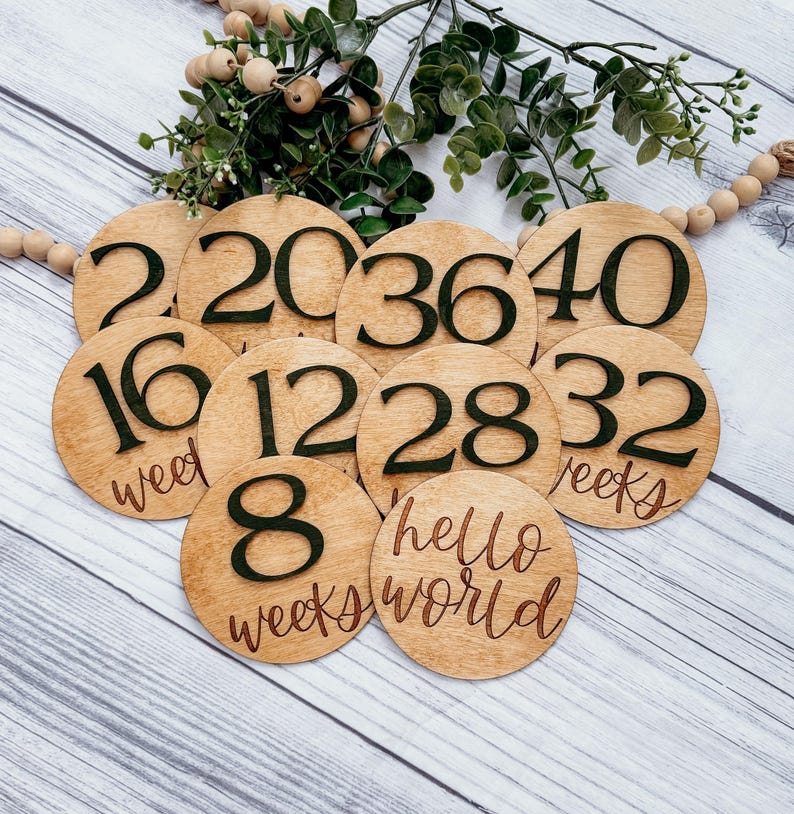 May include: Wooden baby milestone discs with black numbers and text. The discs include the numbers 8, 12, 16, 20, 28, 32, 36, 40, and the text "weeks" and "hello world".