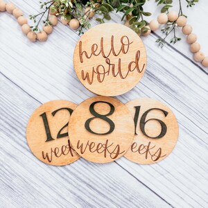 May include: Three wooden circles with the words "hello world", "12 weeks", "8 weeks", and "16 weeks" written on them. The circles are arranged in a triangle shape on a white wooden background.