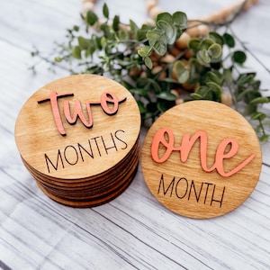 May include: Two wooden baby milestone discs with pink lettering. The first disc says "two months" and the second disc says "one month".