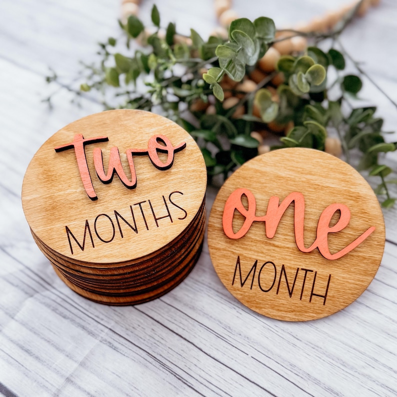 May include: Two wooden baby milestone discs with pink lettering. The first disc says "two months" and the second disc says "one month".