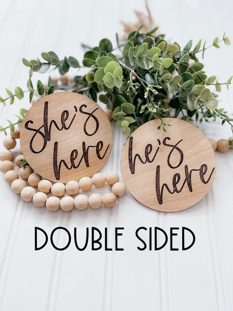 Puede incluir: Dos c&iacute;rculos de madera con el texto "She's here" y "He's here" en una fuente cursiva. Los c&iacute;rculos est&aacute;n sobre una superficie blanca con una hebra de cuentas de madera y una rama de vegetaci&oacute;n.