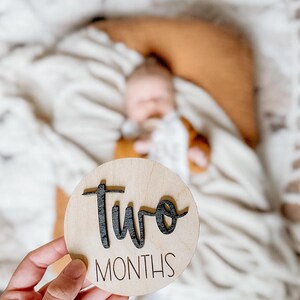 May include: A round wooden sign with the words "two months" written in black lettering. The sign is being held in a person's hand.