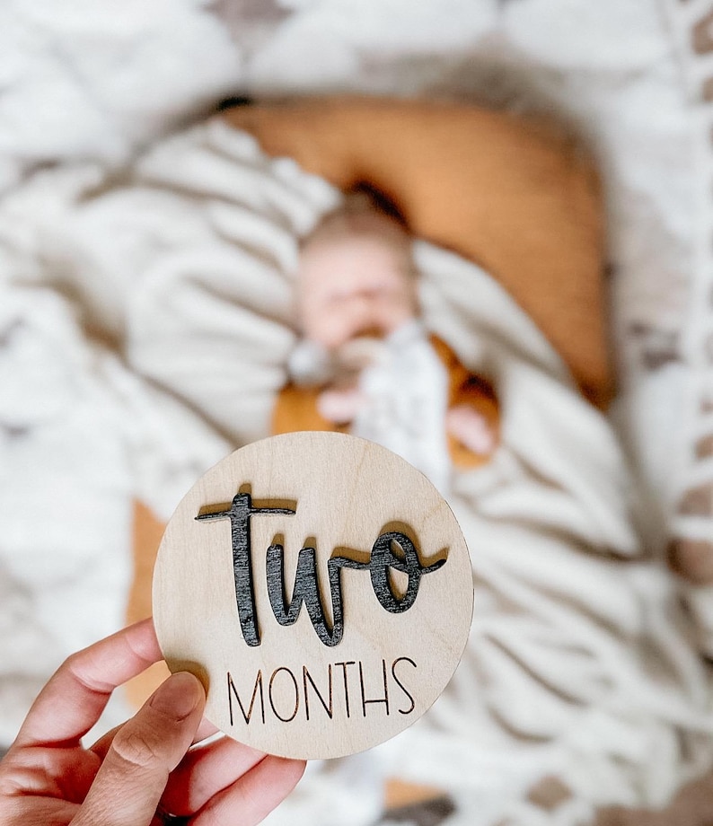 May include: A round wooden sign with the words "two months" written in black lettering. The sign is being held in a person's hand.