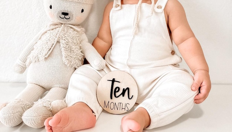 May include: A baby wearing a white ribbed jumpsuit and holding a wooden sign that says "Ten Months". A white stuffed animal is sitting next to the baby.