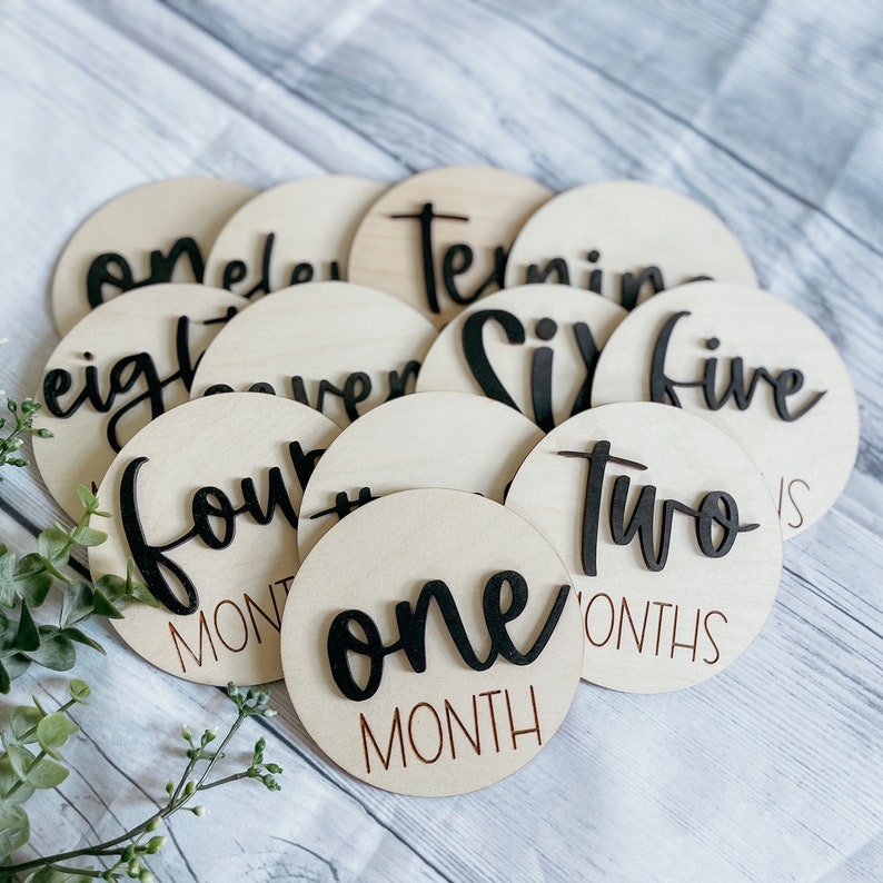 May include: A set of 12 wooden baby milestone discs with black script numbers and the words "month" or "months". The discs are arranged in a circle with the number "one" in the center.