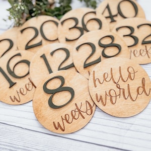 May include: Wooden circles with black numbers and text, showing the progression of weeks during pregnancy. The numbers include 8, 12, 16, 20, 28, 32, 36, 40. One circle says "hello world" and another says "8 weeks".