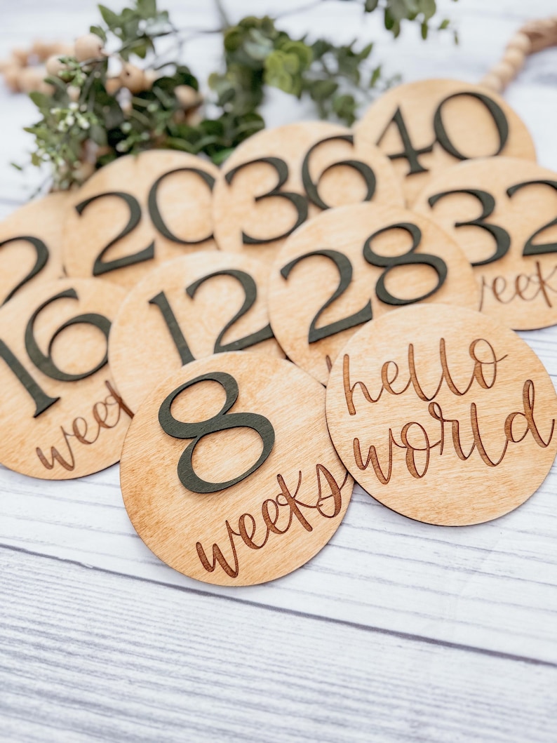 May include: Wooden circles with black numbers and text, showing the progression of weeks during pregnancy. The numbers include 8, 12, 16, 20, 28, 32, 36, 40. One circle says "hello world" and another says "8 weeks".