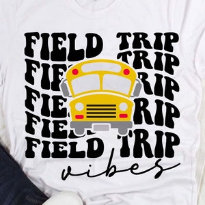 May include: White t-shirt featuring a yellow school bus graphic with the words "FIELD TRIP" in a repeating pattern. Below the bus, the word "vibes" is written in a script font. The shirt is casual wear.
