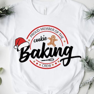 May include: White t-shirt with a red and white Santa hat, a gingerbread man, and a whisk. The text reads "Proud Member of the Cookie Baking Crew".