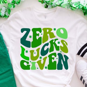 May include: White t-shirt with a green and white graphic that says "Zero Lucks Given" in a retro font. The graphic is surrounded by shamrocks.