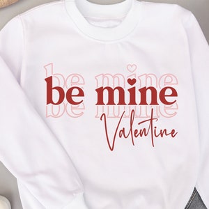 May include: White sweatshirt with the text "be mine Valentine" in red and pink lettering. The text "be" is stacked on top of "mine" with a small heart symbol between the words. The text "Valentine" is written in cursive below the other text.