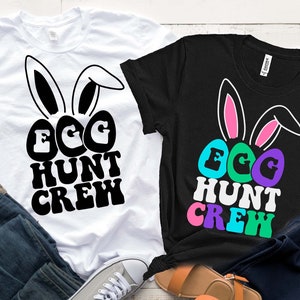 Puede incluir: Dos camisetas con el texto "Egg Hunt Crew" en una fuente retro. Una camiseta es blanca con texto negro y la otra es negra con texto de colores. Ambas camisetas tienen orejas de conejo con el interior rosa.
