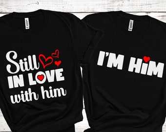 May include: Two black t-shirts with white text and red heart accents. One shirt reads "Still IN LOVE with him," and the other says "I'M Him." The shirts are likely for couples or partners.