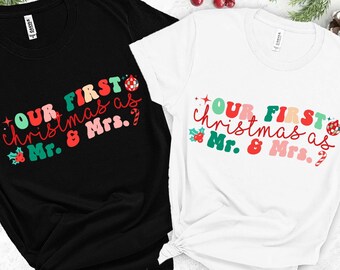 May include: Two t-shirts, one black and one white, featuring the text "OUR FIRST Christmas as Mr. & Mrs." in a colourful, retro font. The design includes festive holly and bauble graphics, ideal for a couple's first Christmas.