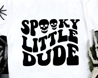 May include: White t-shirt with the words "Spooky Little Dude" in black, stylized font. The design includes two skull graphics. Decorative black spiders and a skeleton hand are placed around the shirt.