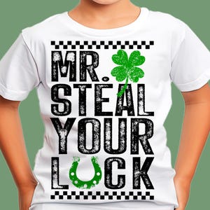 Pode incluir: Camiseta branca com o texto "MR. STEAL YOUR LUCK" em preto, um trevo verde de quatro folhas e uma ferradura verde. A parte superior e inferior da camiseta têm um padrão xadrez em preto e branco.
