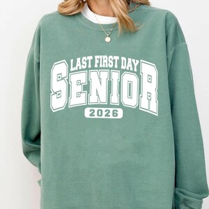 May include: A sage green sweatshirt featuring the text "LAST FIRST DAY SENIOR 2026" in white, block lettering. The sweatshirt has a crew neck and long sleeves. The text is in a collegiate style font.