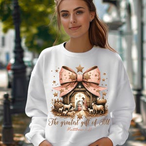 May include: White sweatshirt with a pink bow and a nativity scene with the text "The greatest gift of all Matthew 1:21".