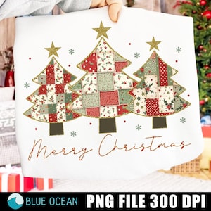 May include: White sweatshirt featuring three Christmas trees with patchwork patterns in red, green, and white, topped with gold stars. The phrase "Merry Christmas" is written in a brown script below the trees. Snowflakes are scattered around the trees.