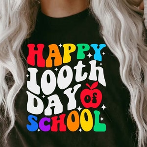以下が含まれることがあります： 「Happy 100th Day of School」と虹色のグラフィックが書かれた黒いTシャツ。中央には赤いリンゴがあります。