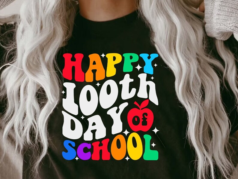 以下が含まれることがあります： 「Happy 100th Day of School」と虹色のグラフィックが書かれた黒いTシャツ。中央には赤いリンゴがあります。