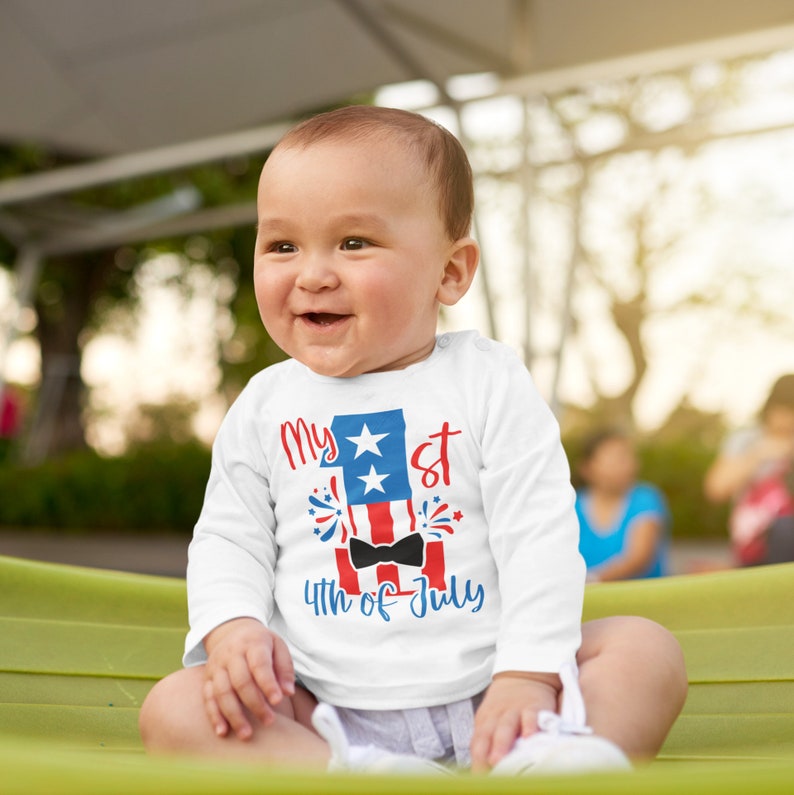 May include: A white long-sleeve baby onesie with a red, white, and blue graphic of a number one with stars and fireworks. The graphic also includes a bow tie and the text "My 1st 4th of July".