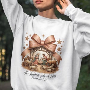 May include: White sweatshirt with a gold and brown illustration of a nativity scene with a bow and stars. The text "The greatest gift of all Matthew 1:21" is printed below the illustration.