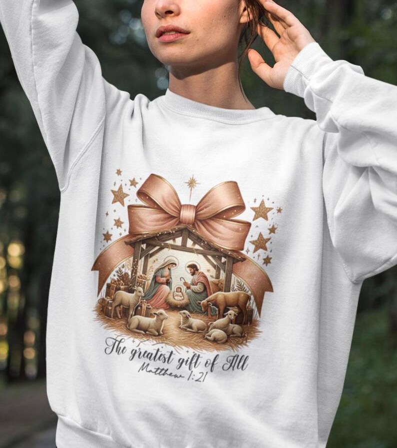 May include: White sweatshirt with a gold and brown illustration of a nativity scene with a bow and stars. The text "The greatest gift of all Matthew 1:21" is printed below the illustration.