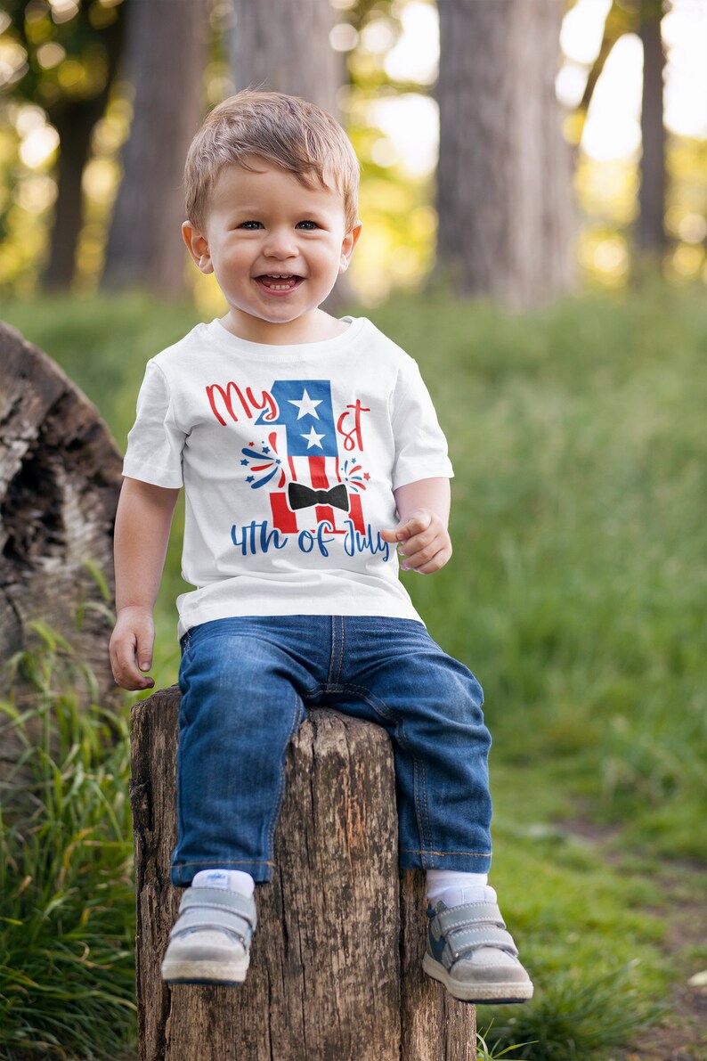 May include: A toddler wearing a white t-shirt with a red, white, and blue graphic that says "My 1st 4th of July" with a bow tie and fireworks. The toddler is sitting on a tree stump in a grassy area.