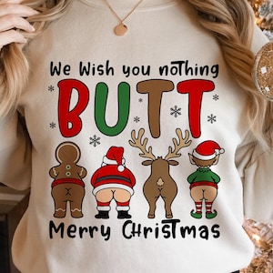 Peut inclure: Sweat-shirt beige avec l'inscription "We Wish You Nothing BUTT Merry Christmas". Le motif présente un bonhomme en pain d'épices, le Père Noël, un renne et un elfe, chacun avec son postérieur exposé. Le texte et les graphiques sont de couleurs variées.