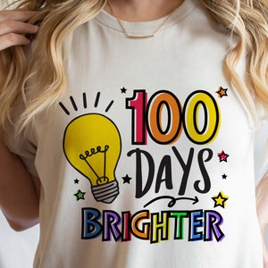 Peut inclure: T-shirt blanc avec un graphique color&eacute; qui dit "100 jours plus lumineux". Le graphique pr&eacute;sente une ampoule, des &eacute;toiles et une fl&egrave;che arc-en-ciel.