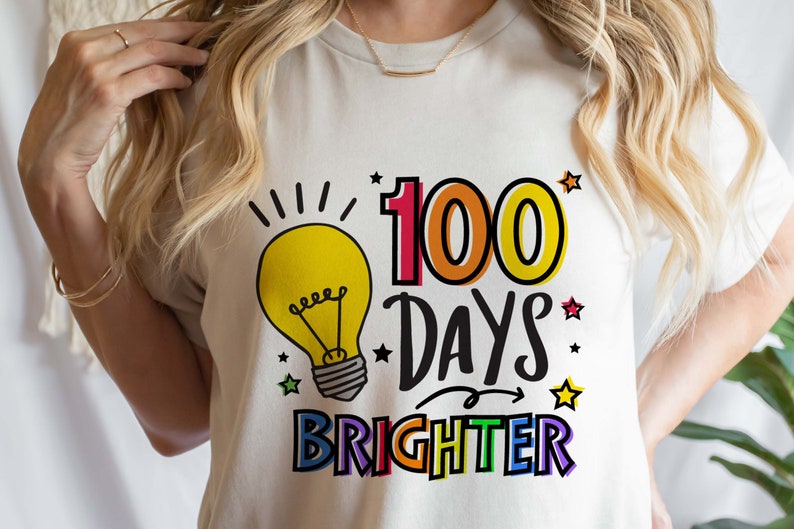 Peut inclure: T-shirt blanc avec un graphique color&eacute; qui dit "100 jours plus lumineux". Le graphique pr&eacute;sente une ampoule, des &eacute;toiles et une fl&egrave;che arc-en-ciel.