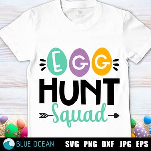 Puede incluir: Camiseta blanca con las palabras "EGG HUNT Squad" en negro, turquesa y blanco. La palabra "EGG" está escrita dentro de tres formas de huevo coloridas. La camiseta es de manga corta y cuello redondo.